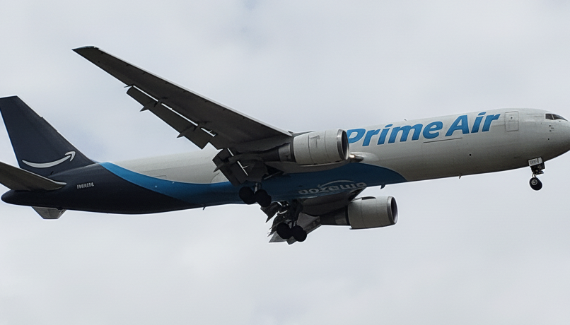 An Amazon Prime Air aircraft flying with its landing gear lowered against a cloudy sky.
