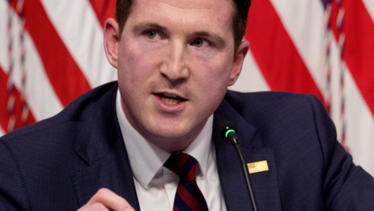 Colin M McDonald speaks into a microphone with American flags behind him.