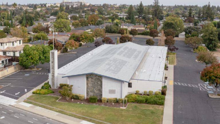 An elevated view of a church building with a pale roof, a tower, surrounding parking areas and nearby homes.