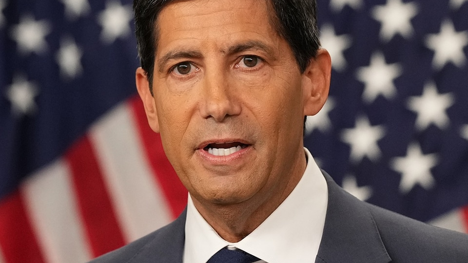 A man in a dark suit and tie speaks with U.S. flags behind him.