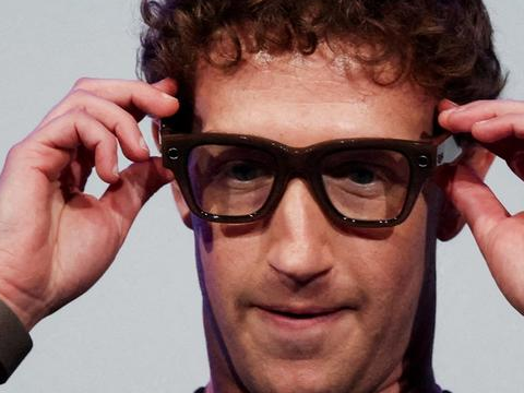 Mark Zuckerberg adjusts thick black glasses with both hands in a close-up portrait.