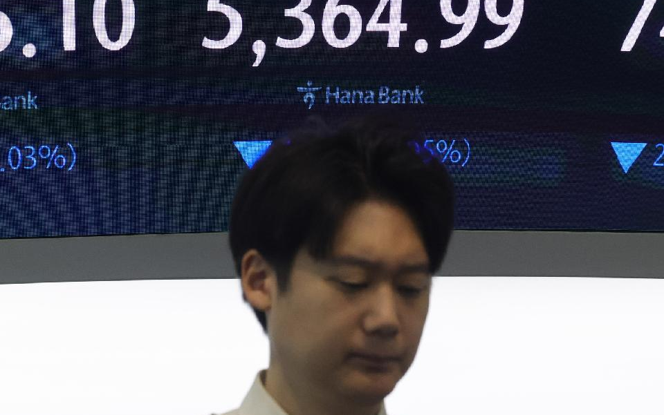 금융시장 전광판 앞에 있는 사람. 뒤로 지수 수치와 파란색 하락 표시가 보인다.