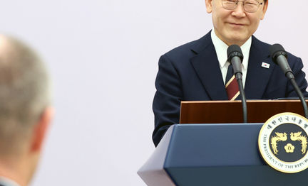 President Lee Jae-myung at a lectern bearing the presidential emblem, with an attendee seen from behind on the left.