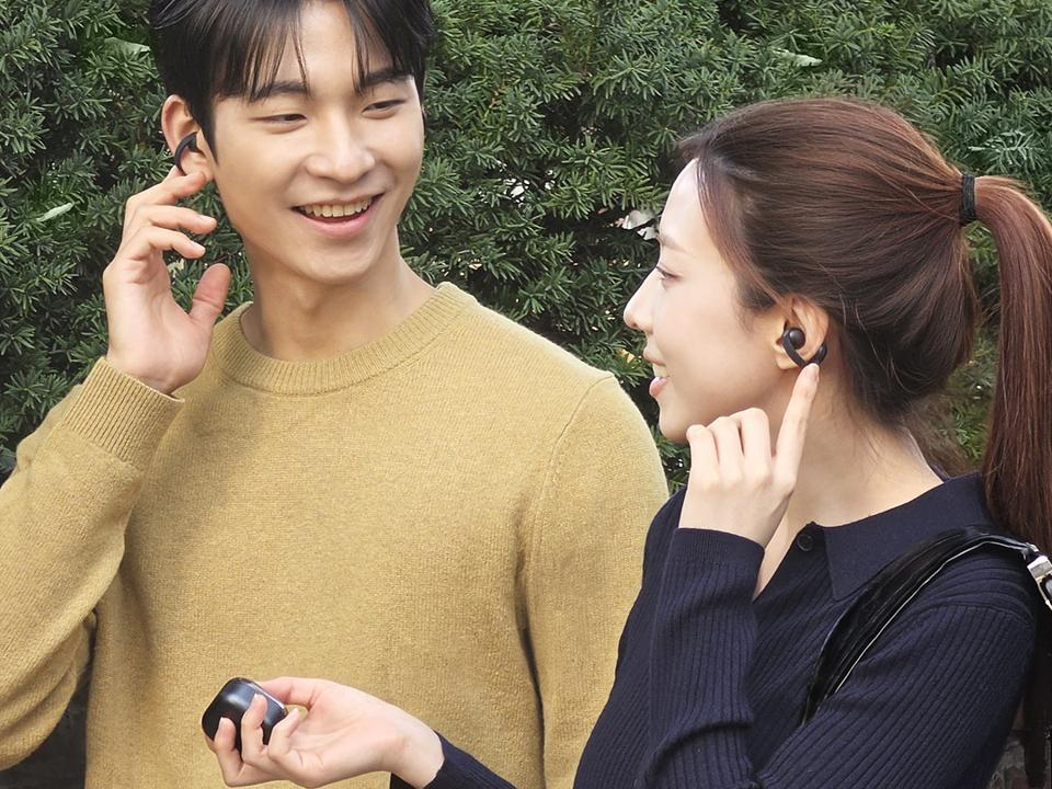 Two people outdoors wearing black clip-style earbuds point to the earbuds, while one holds a black charging case.