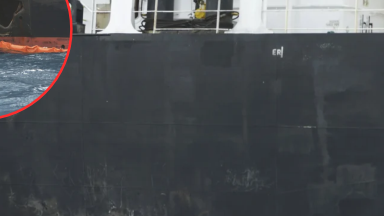Close view of a dark ship hull with surface marks and a partially cropped circular inset showing hull damage near the water.