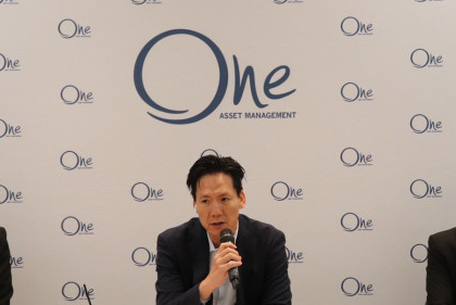 A seated man holds a microphone in front of One Asset Management branding.