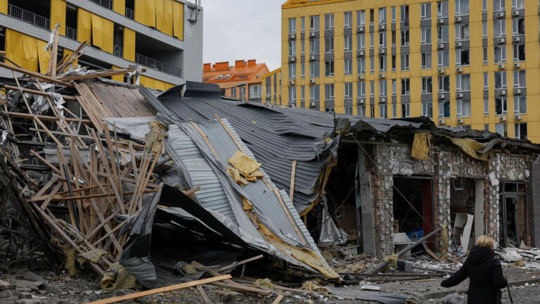 A person walks past debris beside a damaged building with a collapsed roof.