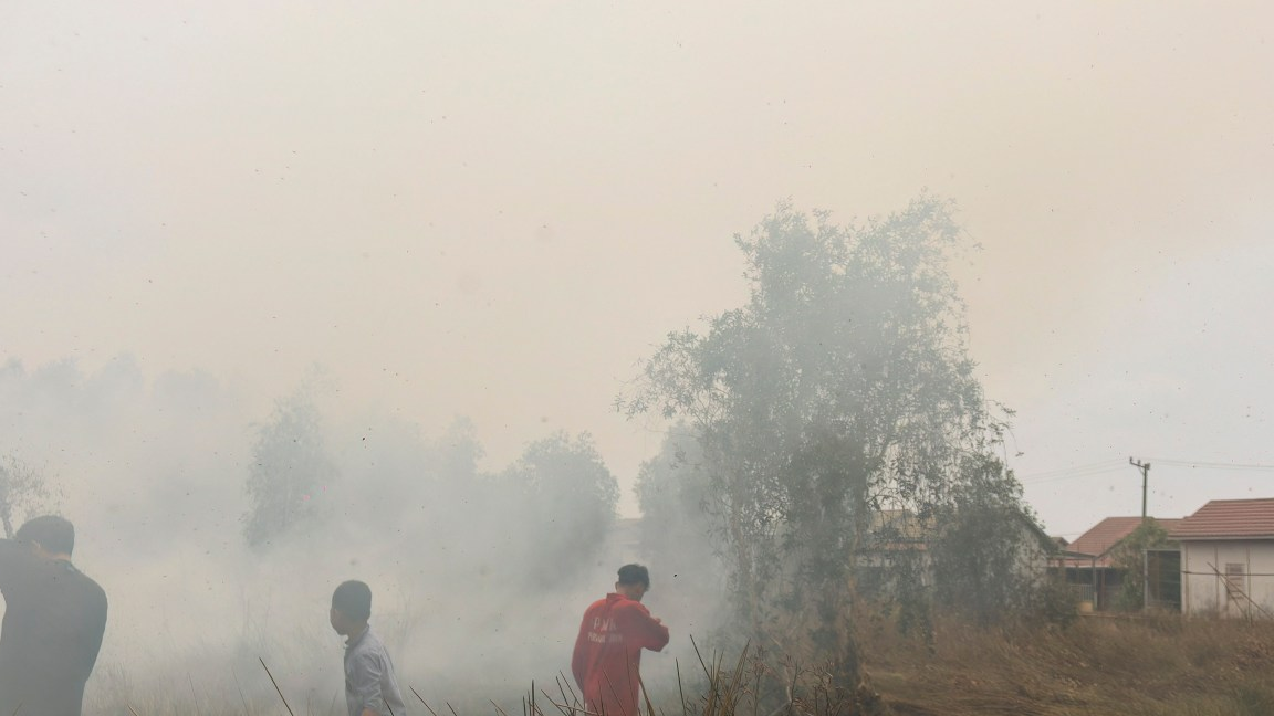 Three people amid thick smoke and dry vegetation, with houses visible on the right.