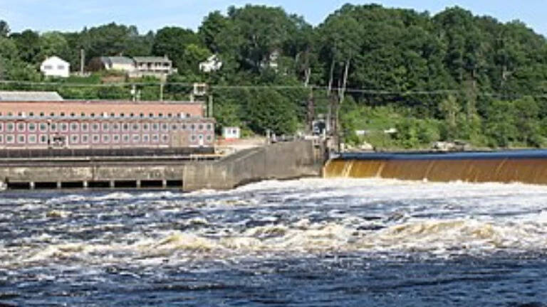 Water flows over a low dam beside a long riverside building and trees.
