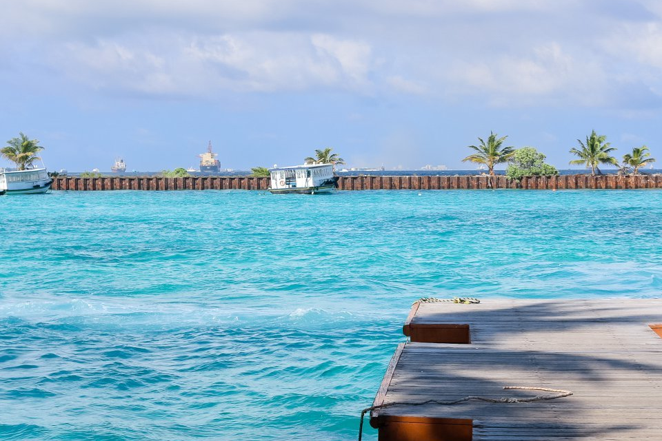 Coastal scenery with turquoise water, palm trees and boats in the distance, and a wooden pier in the right foreground