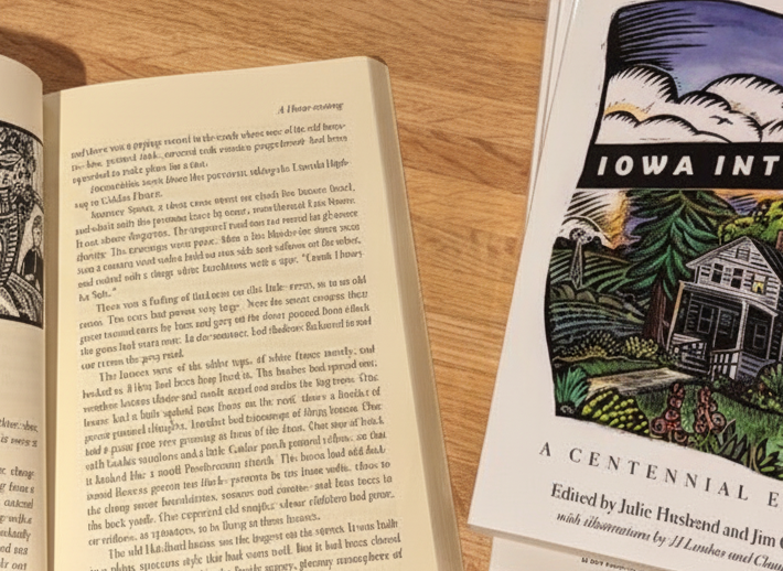 An open book beside stacked copies of Iowa Interiors on a wooden table.