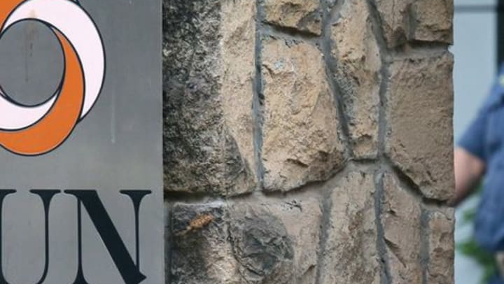 Cropped view of part of a Sun Pharma sign on a stone wall, with a uniformed guard partly visible at the right.