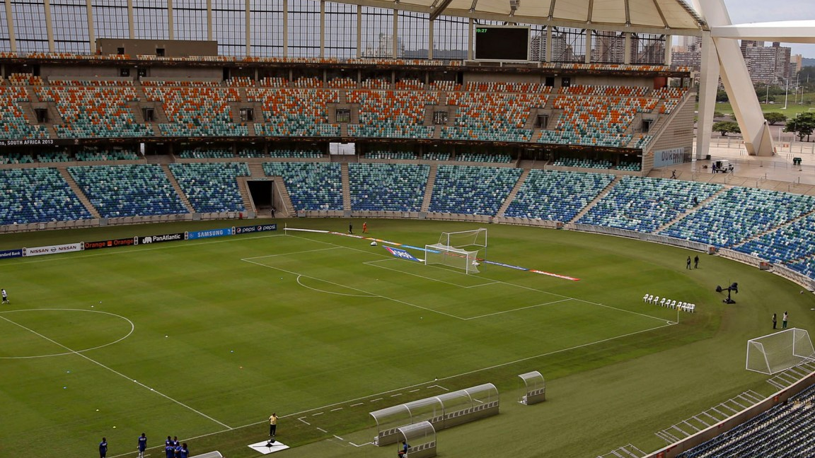 A stadium interior with a grass pitch, multicolored seating and part of its roof structure.