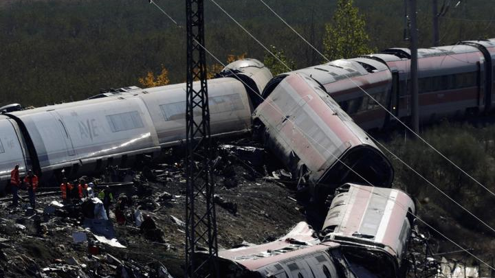 Damaged passenger train carriages lie across railway tracks near emergency personnel.