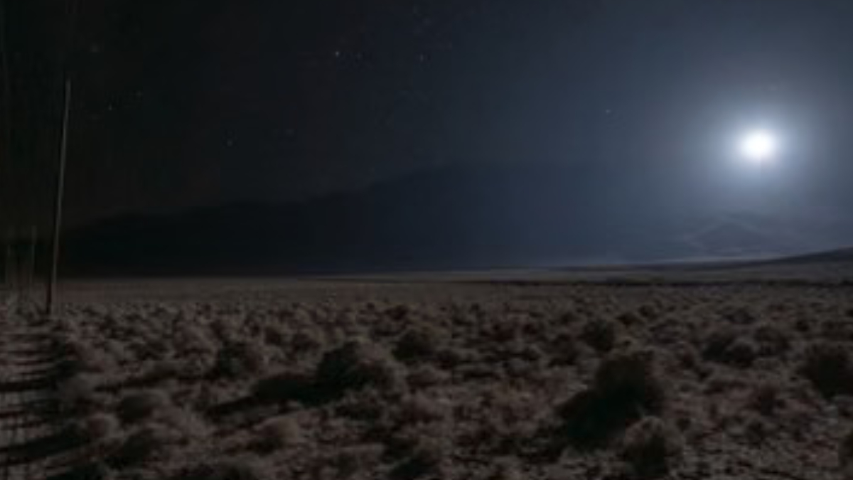 A bright light glows above dark scrubland, with utility poles on the left and distant hills along the horizon.