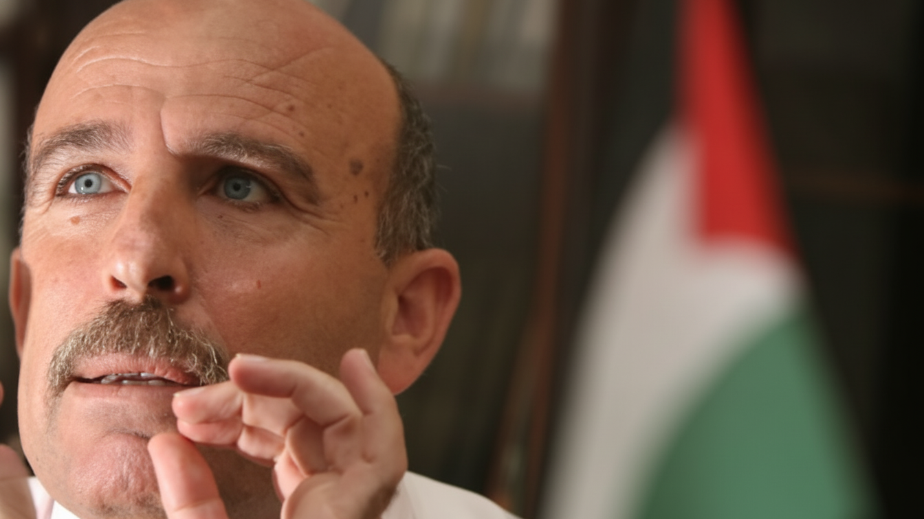 Sufian Abu Zaida gesturing with both hands, with Palestinian flags behind him.