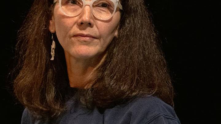 A woman wearing clear-framed glasses and a navy shirt against a dark background.