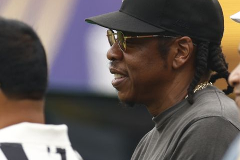 Jay-Z wearing a black cap and sunglasses among a group of people.