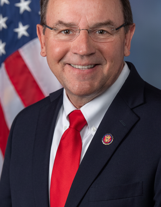 Tom Tiffany wearing glasses, a dark suit and a red tie, with a U.S. flag behind him