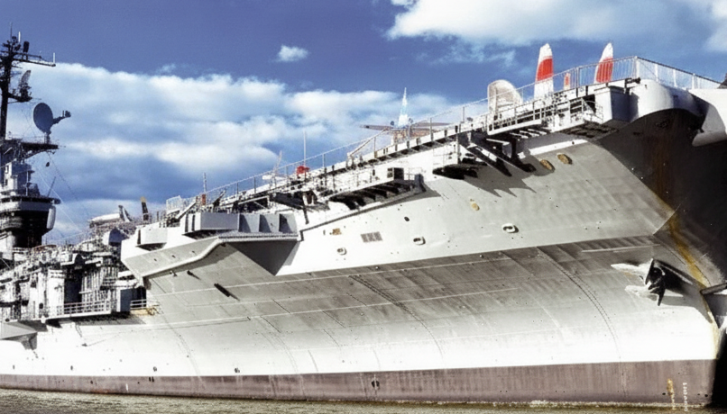 The gray hull and flight deck of the aircraft carrier Intrepid beside the water.