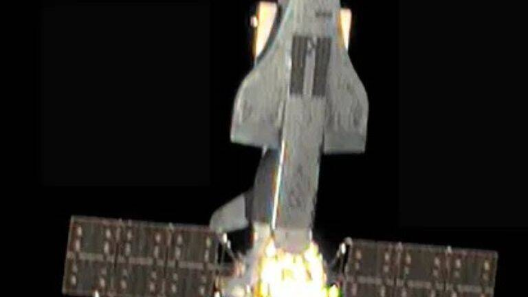 Cropped view of a winged spacecraft with solar panels extending to both sides against a dark background.