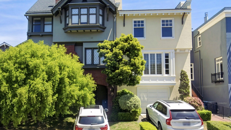 A multi-story house with bay windows and a garage, partially screened by trees, with two cars parked in front.