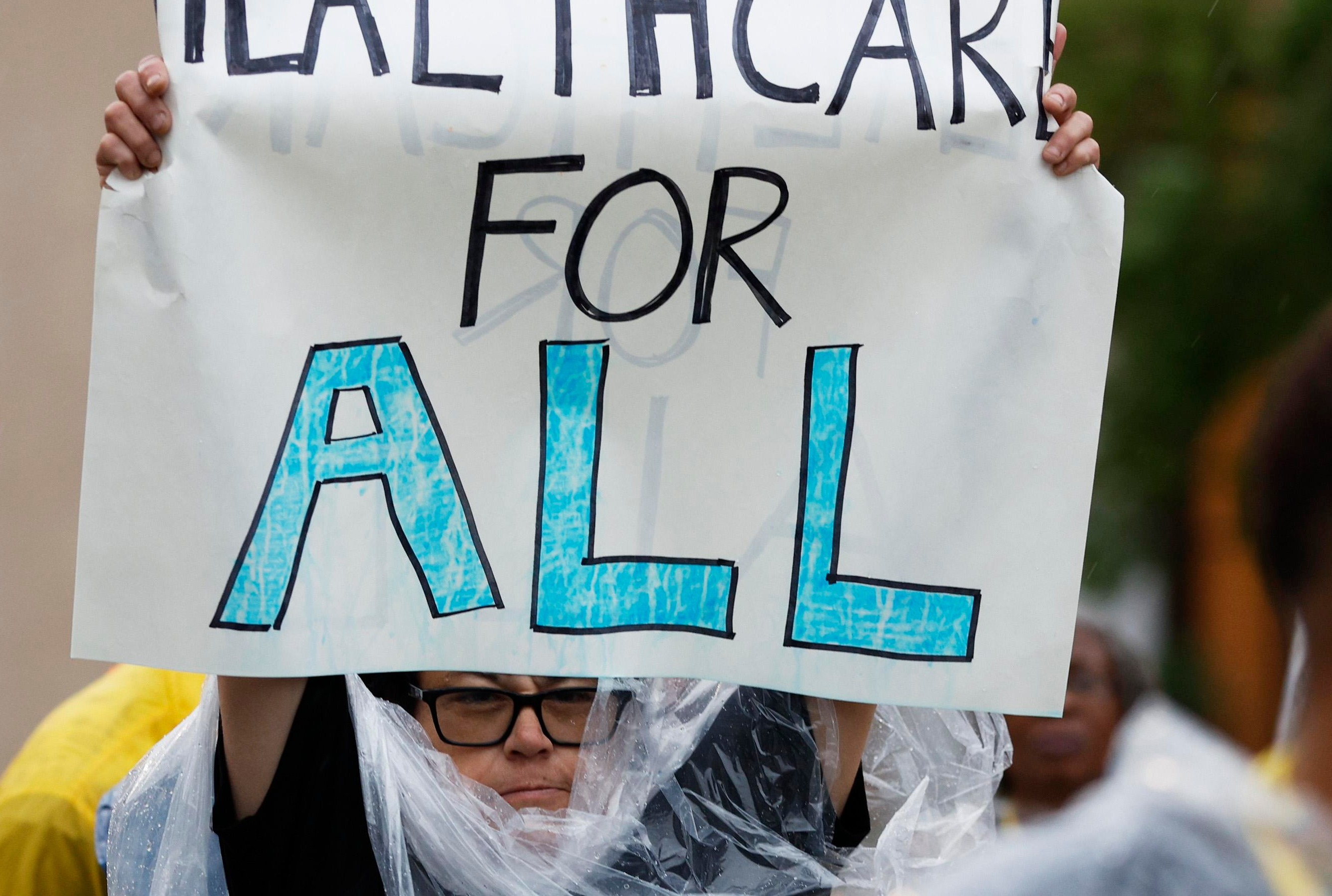 A person wearing glasses and a clear rain poncho holds a handwritten Healthcare for All sign, with its upper edge cropped.